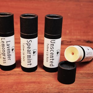 May include: Four lip balm tubes on a wooden surface. Three are upright, labeled "Lavender Lemongrass", "Spearmint", and "Unscented". One tube is open, revealing the balm. The tubes have black caps and white labels.