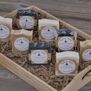 May include: A wooden tray filled with various handmade soap bars. The soaps are labeled with the brand "Tallow Time" and feature different scents like lavender, charcoal, and sweet orange. The soaps are arranged on a bed of shredded paper.