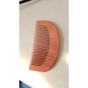Puede incluir: Un peine de madera con un mango curvo y dientes muy juntos. El peine es de madera de color marrón claro y está diseñado para peinar el cabello. El peine está sobre una superficie blanca.