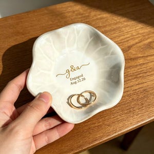 May include: A white ceramic ring dish with a floral design and gold text that reads "g&a" and "Engaged Aug.12.26". Two gold wedding rings rest inside the dish. The dish is held in a hand.