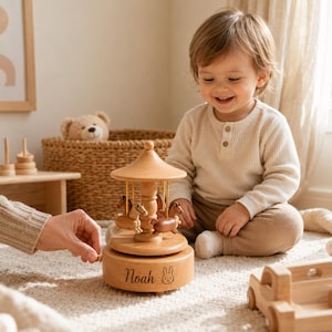 May include: A light brown wooden carousel music box featuring small carved horses suspended under a conical roof. The base is engraved with the name Noah and a small rabbit icon. A hand is shown turning the winding mechanism on the side.