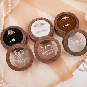 Könnte beinhalten: Runde Holz-Ringboxen mit transparenten Deckeln und Samt-Innenräumen. Einige Boxen enthalten Ringe mit Edelsteinen. Eine Box hat den Text "S & T 09.16.2025". Eine andere Box hat den Text "Mr. & Mrs Frank 09.15.25".