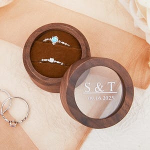 May include: A wooden ring box with a clear lid, engraved with "S & T" and "09.16.2025". Inside, two rings rest on a brown velvet lining. Two additional rings are visible to the left. The box is open, revealing the rings.