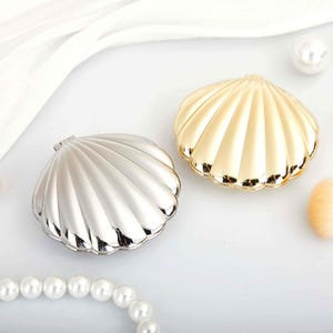 May include: Two compact mirrors shaped like seashells, one with a polished silver finish and the other with a polished gold finish. Both items feature a ribbed, fan-like texture and a hinged design, presented alongside a string of white pearls.