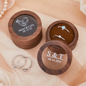 May include: Wooden ring boxes with engraved text and two rings. One box is open, revealing two rings on a brown velvet interior. The other boxes have clear tops with engraved names and dates. The rings are silver with small gemstones.