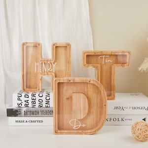 Könnte beinhalten: Drei Buchstaben-Spardosen aus Holz, die "HDT" buchstabieren, mit den Namen "Henry", "Do" und "Tin" darauf geschrieben. Die Spardosen sind aus hellem Holz gefertigt und haben transparente Kunststofffronten. Sie werden auf einer weißen Oberfläche ausgestellt.