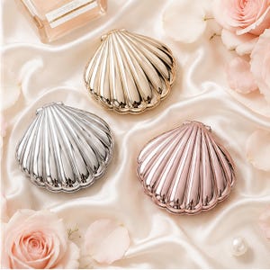 May include: Three compact mirrors shaped like seashells in gold, silver, and rose gold metallic finishes. Each mirror features a hinged design with a ribbed, scalloped exterior surface, arranged on a soft, draped fabric with light pink rose petals.