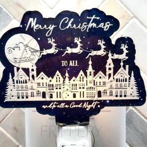 May include: A white night light plug-in with a blue and white design featuring a Christmas scene with the text "Merry Christmas to all and to all a good night."