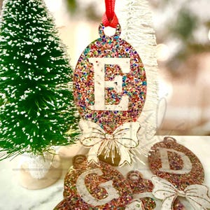 May include: A set of colorful glitter Christmas ornaments with white bows and letters. The ornaments are in the shape of a circle and have a red ribbon attached to the top. The letters on the ornaments are E, G, and D.