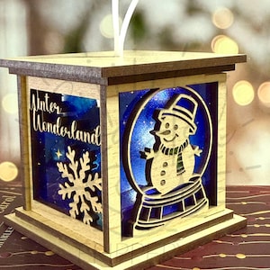 May include: Wooden Christmas lantern with a snowman inside a snow globe design. The lantern has a dark brown top and base, with the words "Winter Wonderland" and a snowflake on one side. The text "DIGITAL FILE ONLY" is at the top.