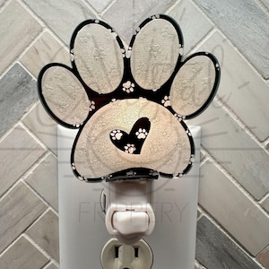 Puede incluir: Una luz nocturna blanca con forma de huella de pata con un contorno negro y huellas de pata blancas en el interior. La huella de pata tiene un corazón en el centro con huellas de pata blancas en el interior. La luz nocturna está enchufada a una toma de corriente estándar.