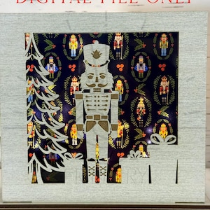 May include: A wooden Christmas-themed shadow box with a light gray frame. The interior features a cutout of a nutcracker, Christmas tree, and presents against a navy blue background with nutcracker illustrations. The text "DIGITAL FILE ONLY" is at the top.