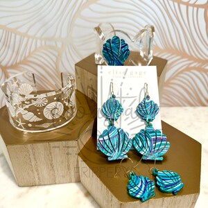May include: Teal and purple seashell earrings with gold hooks. The earrings are displayed on a wooden stand with the text "Elise Gage Coast &  Whimsy" on a white card.