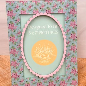 May include: A rectangular picture frame with a floral pattern in pink and blue. The frame has a pink scalloped oval opening for a 5 x 7 inch picture. The frame has the text "Designed To Fit 5 x 7" PICTURES". The frame also has a circular logo.