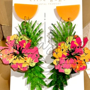 May include: A pair of earrings featuring a tropical flower design with green leaves and a half-circle orange accent. The earrings are made of acrylic and have a gold-colored metal hook.