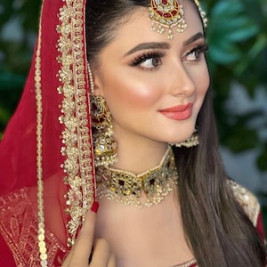 May include: A woman wearing a red bridal outfit with gold embroidery and a matching veil. She is wearing a gold and red jeweled necklace and earrings. She has a red bindi on her forehead.