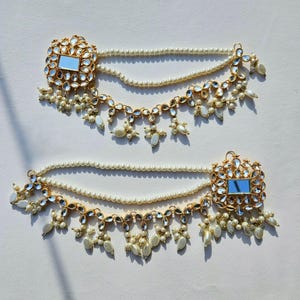 Könnte beinhalten: Ein Paar goldene und weiße Perlen-Haarketten mit einem Spiegeldetail. Jede Kette hat einen quadratischen Anhänger mit einem kleinen Spiegel in der Mitte, umgeben von klaren Steinen. Die Ketten bestehen aus kleinen weißen Perlen und haben ein baumelndes Design mit mehreren Perlentropfen.