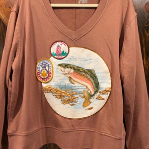 May include: A dusty rose V-neck sweatshirt featuring a large circular graphic of a trout jumping from water. Two embroidered patches are attached: a round Scout-O-Rama patch and an oval Boypower Land O'Lakes 1969 patch. Terra & Sky label.