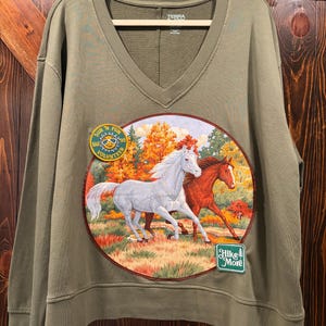 May include: An olive green long-sleeve sweatshirt with a V-neckline. The front features a circular graphic of a white horse and a brown horse running through an autumn forest. It includes a yellow circular patch reading Sun 'N Fun Volunteer and a green rectangular patch with the text Hike More.