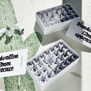 May include: White stamp sets with black lettering, including uppercase and lowercase options, are displayed. The text "Handwritten 12mm" is visible on the white labels. The stamps are in white boxes.