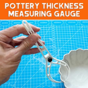 Könnte beinhalten: Weißes Messgerät zur Messung der Dicke von Keramik mit dem Text "POTTERY THICKNESS MEASURING GAUGE". Das Messgerät wird über einer weißen Keramikschale gehalten. Der Hintergrund ist eine blaue Schneidematte mit Messungen.