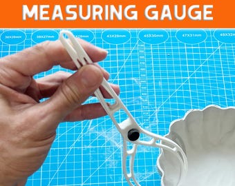 May include: A white pottery thickness measuring gauge with the text "POTTERY THICKNESS MEASURING GAUGE". The gauge is held over a white ceramic bowl. The background is a blue cutting mat with measurements.
