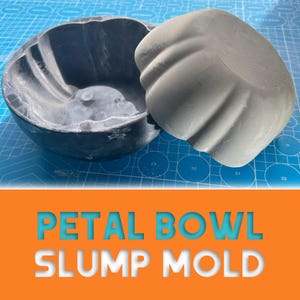Op de afbeelding: Een grijze petal bowl slump mold, met een bijpassend deksel, ligt op een blauwe snijmat. De mal is ontworpen met een bloembladachtige vorm. De tekst "PETAL BOWL SLUMP MOLD" wordt weergegeven in een witte letter met een blauwe omlijning.