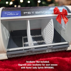 May include: White plastic pet brooder with a clear lid and a red bow. The brooder has a digital control panel and a notice label. The text on the brooder reads "Pet Brooder" and "C.U. RADLE".