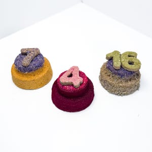 May include: Three small, round, tiered treats with textured surfaces. One is yellow and purple with the number 7, one is magenta with the number 4, and one is brown and purple with the number 16. Each treat features a flat, raised number on top.