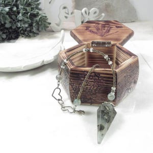 May include: A wooden hexagonal box with an open lid, revealing a necklace with a heart-shaped charm and a crystal pendulum. The box is brown with engraved details. The necklace features beads and a clear, pointed crystal.