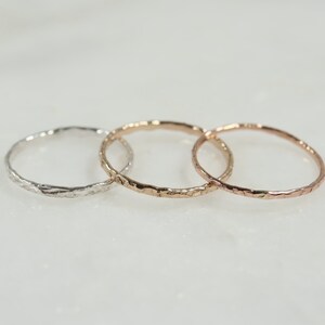 May include: Three delicate rings in silver, rose gold, and gold. Each ring has a textured, hammered finish. The rings are displayed on a white surface, showcasing their different metallic hues and simple design.