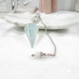 May include: A light blue crystal pendulum with a silver chain and a white, round handle. The crystal is diamond-shaped and faceted. The handle has a silver band with iridescent rhinestones. The pendulum is designed for divination.
