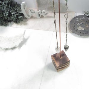 May include: A wooden jewelry display stand with two copper-colored rods holding decorative pendants. The pendants have clear and silver-colored beads and charms. The base is a square block of wood with a dark, burnt finish.