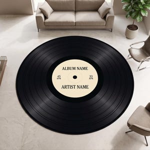 Op de afbeelding: Een groot rond vloerkleed ontworpen als een zwarte vinylplaat. Het centrale label is crèmekleurig met de tekst ALBUM NAME en ARTIST NAME in het zwart gedrukt, samen met de aanduidingen 45 RPM en 33 RPM aan de linker- en rechterkant.