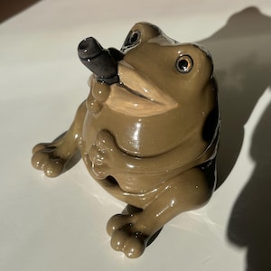 May include: A brown ceramic frog ornament, smoking a black pipe. The frog has large eyes and is in a seated position. The figurine has a glossy finish. The frog is approximately 10 cm in length.