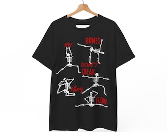 Puede incluir: Camiseta negra con figuras de esqueletos blancos en posturas de yoga. El texto rojo dice "MY BONES DON'T CREAK THEY FLOW". La camiseta está colgada en una percha de madera.