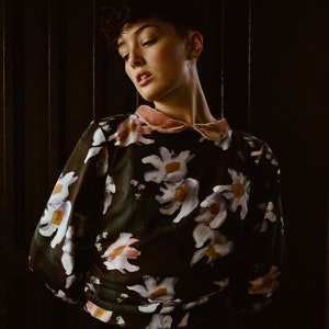 May include: A person with short, dark hair is wearing a black blouse with a pink collar and a floral print. The flowers are white, pink, and purple.