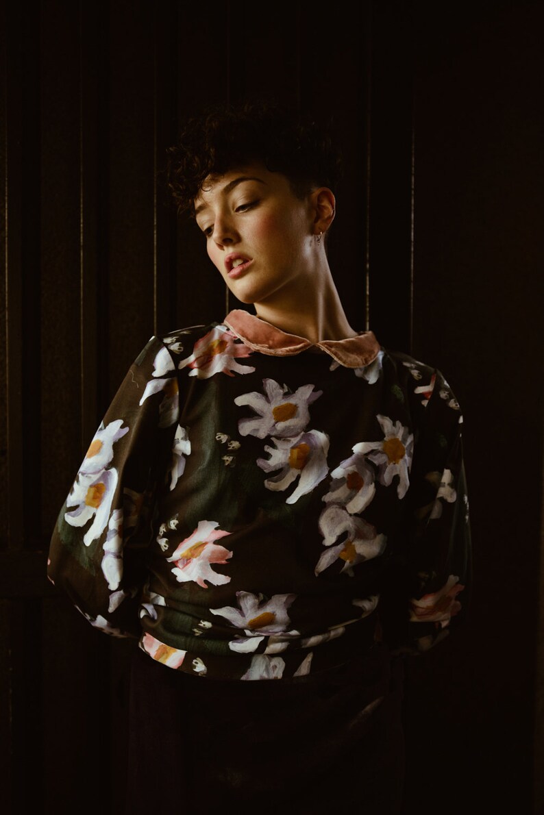 May include: A person with short, dark hair is wearing a black blouse with a pink collar and a floral print. The flowers are white, pink, and purple.