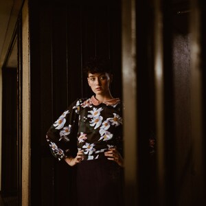May include: A woman with short dark hair stands in a doorway, wearing a black and white floral blouse with a pink collar. She is looking at the camera with a serious expression.