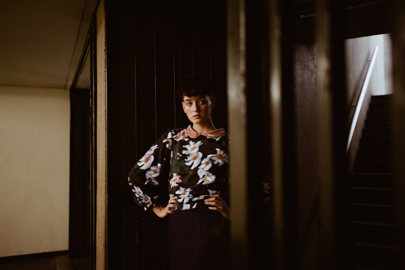 May include: A woman with short dark hair stands in a doorway, wearing a black and white floral blouse with a pink collar. She is looking at the camera with a serious expression.