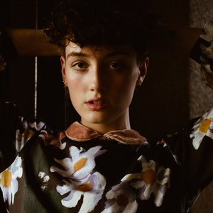May include: A person with short, curly dark hair wearing a black and white floral blouse with a pink velvet collar. The person is looking directly at the camera with their arms behind their head.