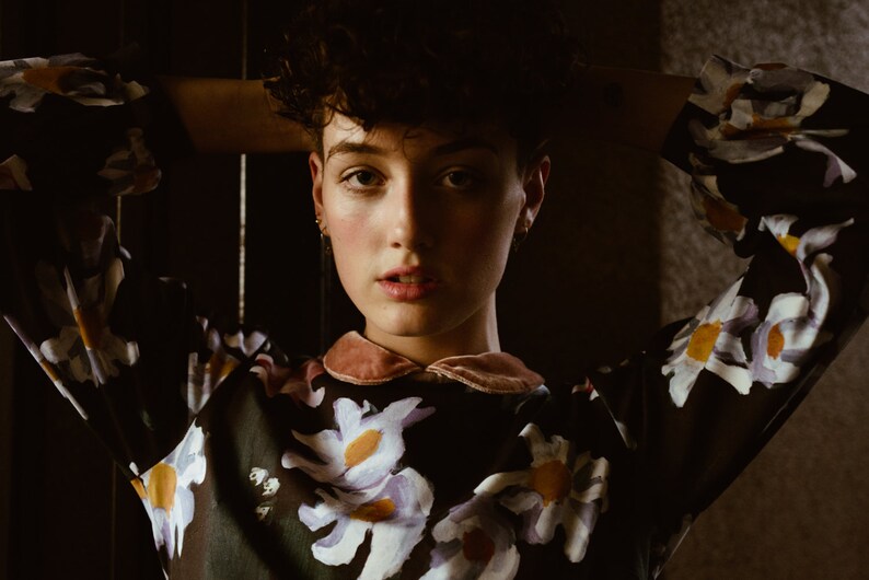 May include: A person with short, curly dark hair wearing a black and white floral blouse with a pink velvet collar. The person is looking directly at the camera with their arms behind their head.