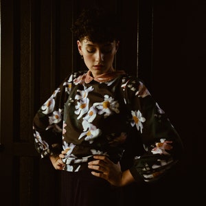 May include: A woman with short dark hair is wearing a black blouse with a floral print. The blouse has a pink collar and long sleeves. She is standing in front of a dark background.