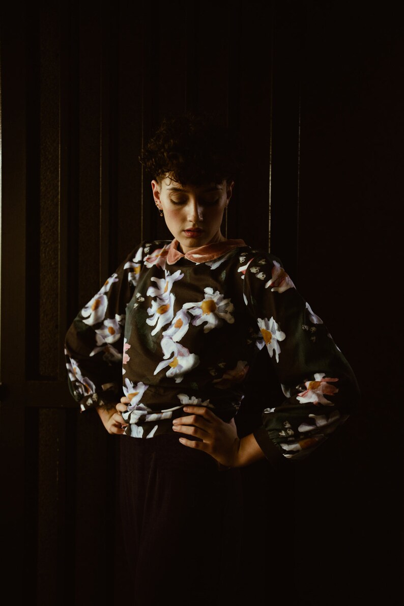 May include: A woman with short dark hair is wearing a black blouse with a floral print. The blouse has a pink collar and long sleeves. She is standing in front of a dark background.