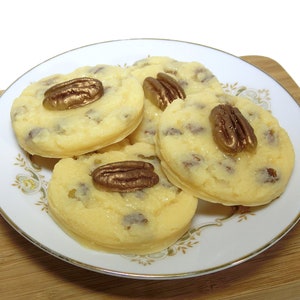 May include: Four pecan-topped cookies on a white plate with a gold trim. The cookies are light yellow and have a smooth, glossy finish. The pecans are a shiny gold color.