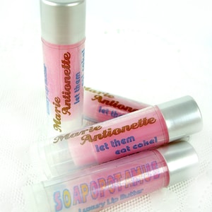 Op de afbeelding: Drie roze lippenbalsem tubes met zilveren doppen. De tubes zijn gelabeld met "Marie Antoinette let them eat cake!" en "SOAPOPOTAMUS Luxury Lip Butter".