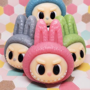 Puede incluir: Una colección de cinco figuras caprichosas, cada una con una cara de dibujos animados con grandes ojos negros y una expresión severa. Las figuras están adornadas con orejas de conejo y vienen en varios colores, incluyendo rosa, azul, gris y verde.