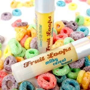 Può includere: Due tubetti di balsamo per le labbra con tappi argentati ed etichette che recitano "Fruit Loops milky cereal bowl" e "Luxury Lip Butter". I tubetti sono circondati da cereali colorati a forma di anello.
