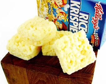 Pode incluir: Quatro quadrados de Rice Krispies em uma tábua de cortar de madeira. As guloseimas são feitas com marshmallows e cereal Rice Krispies. As guloseimas são de cor amarelo claro. Uma caixa de cereal Rice Krispies da Kellogg's está no fundo.
