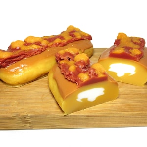 May include: Three glazed donuts with a red and orange bacon topping on a wooden cutting board. The donuts are cut in half to reveal a white filling.